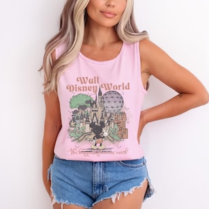 Puede incluir: Una camiseta sin mangas rosa claro con un gr&aacute;fico de Walt Disney World y Mickey Mouse. El texto "Walt Disney World" est&aacute; encima del gr&aacute;fico, y "The happiest place on earth" est&aacute; debajo. La camiseta sin mangas se combina con unos pantalones cortos vaqueros azules.