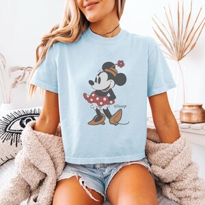 May include: Light blue t-shirt featuring a Minnie Mouse graphic. The cartoon character is depicted in a red polka dot dress, brown shoes, and a matching hat. The word "Disney" is printed below the character.