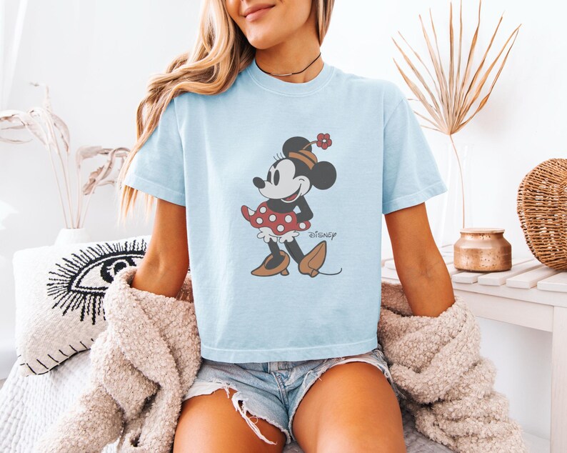 May include: Light blue t-shirt featuring a Minnie Mouse graphic. The cartoon character is depicted in a red polka dot dress, brown shoes, and a matching hat. The word "Disney" is printed below the character.