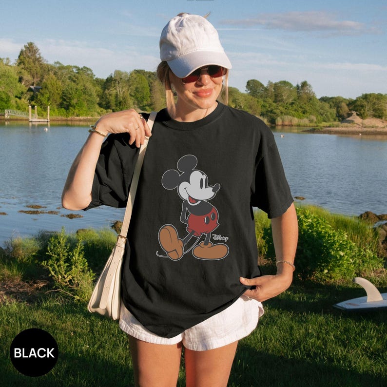 May include: A black short-sleeve t-shirt featuring a graphic print of Mickey Mouse in red shorts and brown shoes, with the Disney logo text. The shirt is paired with a white baseball cap, white shorts, and a beige shoulder bag with a strap.