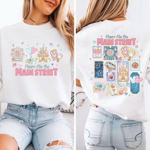 May include: White sweatshirt with a colorful graphic design. The front features a Ferris wheel, castle, popcorn, and balloons, with the text "Meet Me On Main Street." The back has a similar design with stamps.