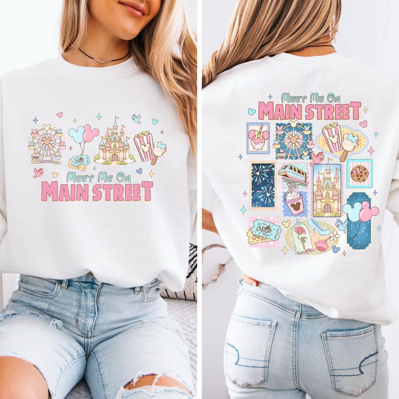 May include: White sweatshirt with a colorful graphic design. The front features a Ferris wheel, castle, popcorn, and balloons, with the text "Meet Me On Main Street." The back has a similar design with stamps.
