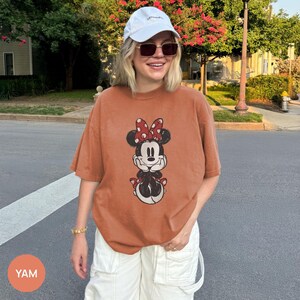 Puede incluir: Una camiseta de color &oacute;xido con un estampado de Minnie Mouse con un lazo rojo de lunares. La camiseta tiene mangas cortas y un corte holgado. La imagen tambi&eacute;n muestra pantalones cargo blancos, una gorra blanca y gafas de sol.