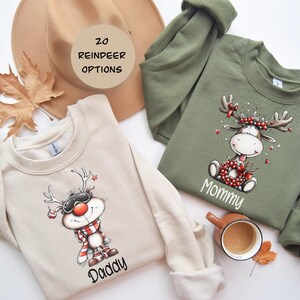 May include: Two sweatshirts with reindeer designs. One is beige with a reindeer wearing goggles and a scarf, the word "Daddy" below. The other is olive green with a reindeer wearing a wreath, with the word "Mommy" below. A hat and leaf are in the background.