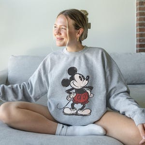 May include: A gray sweatshirt featuring a classic Mickey Mouse graphic in black, red, and white. The sweatshirt is worn by a person sitting on a gray couch. The image is well-lit, showcasing the details of the design.