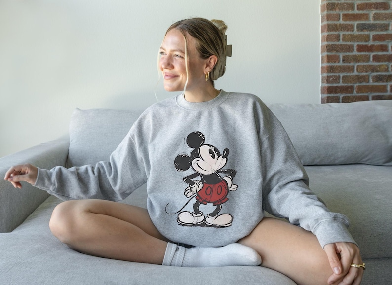 May include: A gray sweatshirt featuring a classic Mickey Mouse graphic in black, red, and white. The sweatshirt is worn by a person sitting on a gray couch. The image is well-lit, showcasing the details of the design.