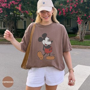 May include: A brown short-sleeve t-shirt featuring a graphic of Mickey Mouse with red shorts and yellow shoes. The shirt is paired with a beige baseball cap and white shorts. A tan shoulder bag strap is visible over the shoulder.