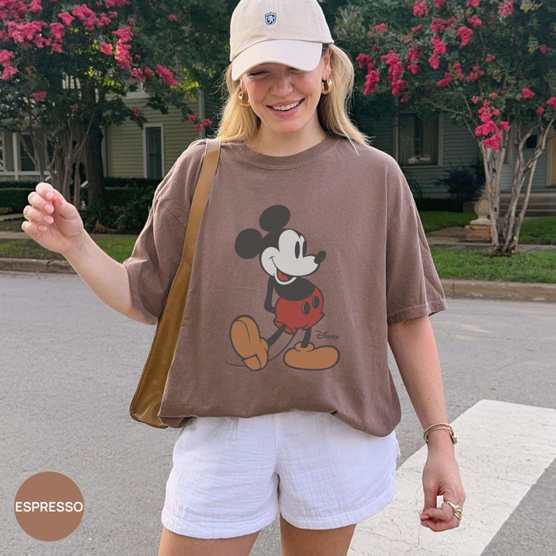 May include: A brown short-sleeve t-shirt featuring a graphic of Mickey Mouse with red shorts and yellow shoes. The shirt is paired with a beige baseball cap and white shorts. A tan shoulder bag strap is visible over the shoulder.