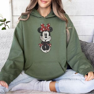 May include: Olive green hooded sweatshirt featuring a graphic of Minnie Mouse. Minnie is depicted in a seated pose, wearing a red and white polka dot bow. The sweatshirt has a front pocket and long sleeves.
