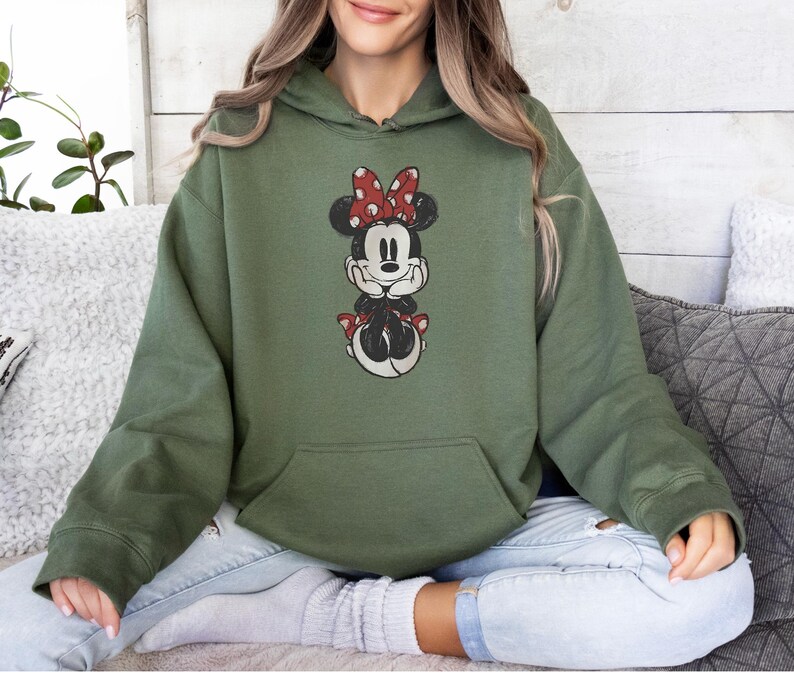 May include: Olive green hooded sweatshirt featuring a graphic of Minnie Mouse. Minnie is depicted in a seated pose, wearing a red and white polka dot bow. The sweatshirt has a front pocket and long sleeves.