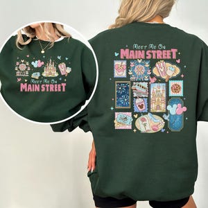 May include: Dark green sweatshirt with a colorful graphic design. The front features a Ferris wheel, castle, popcorn, and text that reads "Meet Me On Main Street." The back has a collage of stamps with various theme park icons.