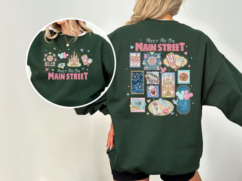 May include: Dark green sweatshirt with a colorful graphic design. The front features a Ferris wheel, castle, popcorn, and text that reads "Meet Me On Main Street." The back has a collage of stamps with various theme park icons.