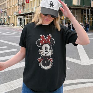 Puede incluir: Camiseta negra con un dise&ntilde;o de Minnie Mouse, lazo rojo y lunares blancos. El conjunto incluye vaqueros azules y una gorra de b&eacute;isbol blanca con el logo "LA" negro. La persona est&aacute; en una calle de la ciudad.