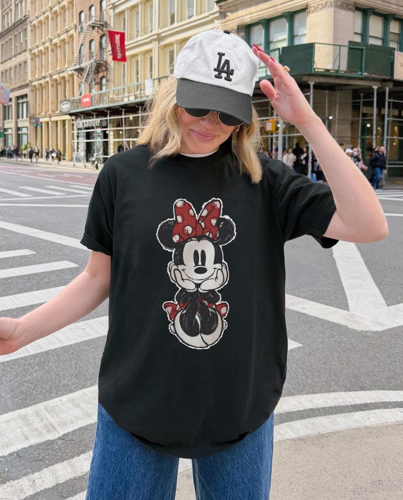 Puede incluir: Camiseta negra con un dise&ntilde;o de Minnie Mouse, lazo rojo y lunares blancos. El conjunto incluye vaqueros azules y una gorra de b&eacute;isbol blanca con el logo "LA" negro. La persona est&aacute; en una calle de la ciudad.