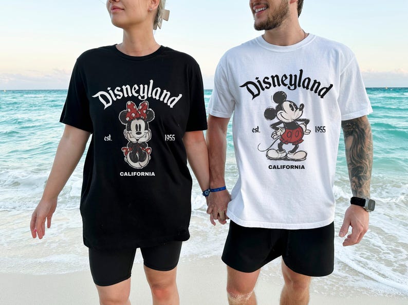 May include: Black and white Disneyland t-shirts featuring Minnie Mouse and Mickey Mouse graphics. The shirts have the Disneyland logo, the text "est. 1955", and "CALIFORNIA". The people are holding hands on a beach.