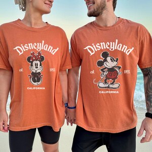May include: Two orange t-shirts with the Disneyland logo and cartoon characters Minnie Mouse and Mickey Mouse. The shirts have the text "est. 1955 CALIFORNIA". The shirts are worn by two people on a beach.