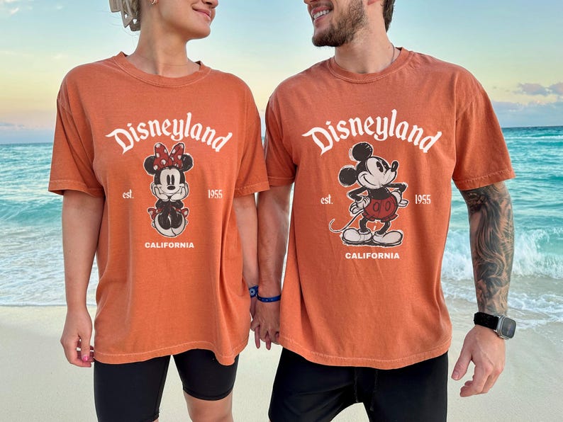 May include: Two orange t-shirts with the Disneyland logo and cartoon characters Minnie Mouse and Mickey Mouse. The shirts have the text "est. 1955 CALIFORNIA". The shirts are worn by two people on a beach.