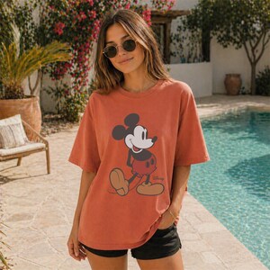 May include: A terracotta-colored short-sleeve t-shirt featuring a graphic print of Mickey Mouse on the front. The character is depicted in a classic pose with black ears, a white face, and red shorts. The word Disney is printed in small black script.