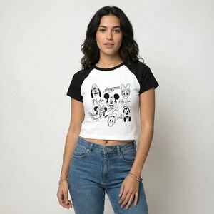 May include: A white and black raglan crop top featuring a graphic of Mickey Mouse and friends. The black graphic includes Pluto, Mickey Mouse, Minnie Mouse, Donald Duck, Daisy Duck, and Goofy. The shirt has black sleeves and a black neckline.
