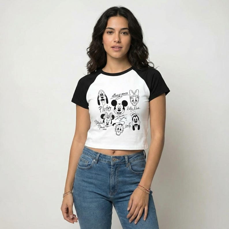 May include: A white and black raglan crop top featuring a graphic of Mickey Mouse and friends. The black graphic includes Pluto, Mickey Mouse, Minnie Mouse, Donald Duck, Daisy Duck, and Goofy. The shirt has black sleeves and a black neckline.