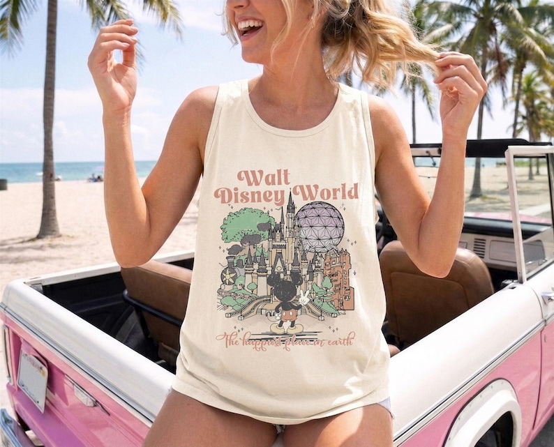 May include: A cream-coloured sleeveless tank top featuring a graphic print of the Walt Disney World castle, Spaceship Earth, and Mickey Mouse. The text reads Walt Disney World and The happiest place on earth in a brown script font.