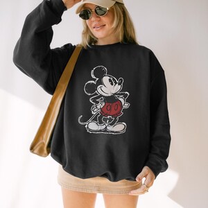 May include: Black sweatshirt featuring a distressed graphic of Mickey Mouse in a classic pose. The cartoon character is depicted in white, red, and black. The sweatshirt is a crew neck style, and the image is centered on the front.
