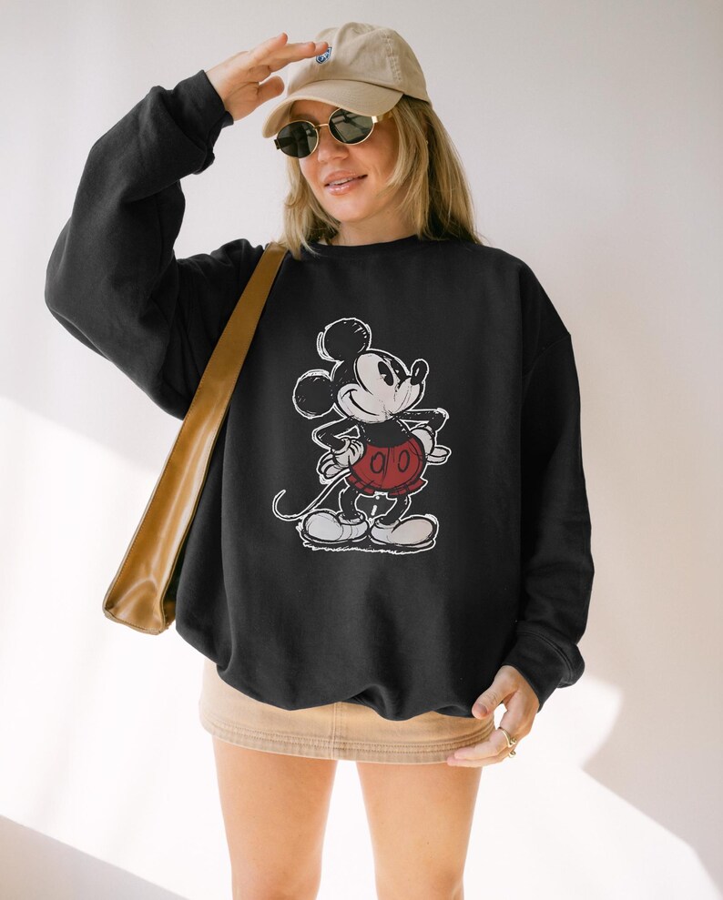 May include: Black sweatshirt featuring a distressed graphic of Mickey Mouse in a classic pose. The cartoon character is depicted in white, red, and black. The sweatshirt is a crew neck style, and the image is centered on the front.