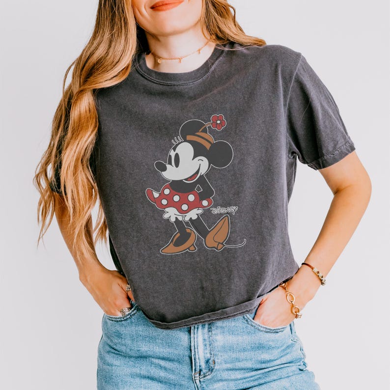May include: Dark gray t-shirt featuring a vintage Minnie Mouse graphic. The cartoon character is depicted in a red and white polka dot dress, brown shoes, and a hat with a red flower. The word "disney" is printed below the graphic.
