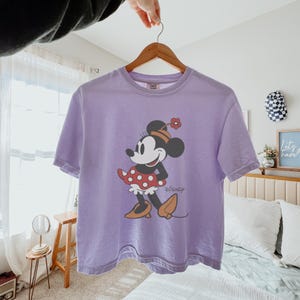May include: A lavender-colored t-shirt featuring a graphic of Minnie Mouse. Minnie is depicted in a red polka-dot skirt, orange shoes, and a flower on her hat. The Disney logo is printed below the character.
