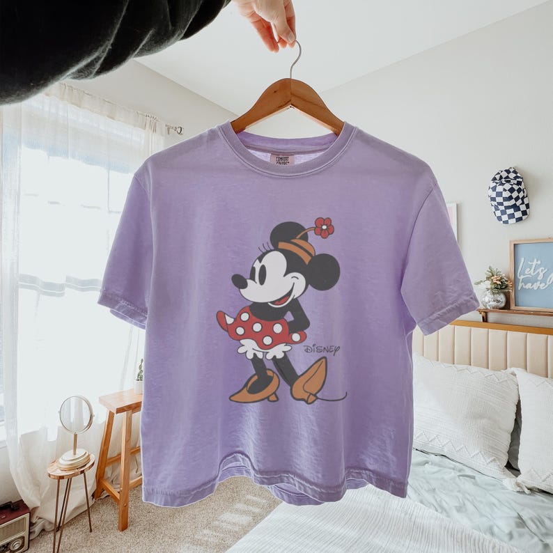 May include: A lavender-colored t-shirt featuring a graphic of Minnie Mouse. Minnie is depicted in a red polka-dot skirt, orange shoes, and a flower on her hat. The Disney logo is printed below the character.