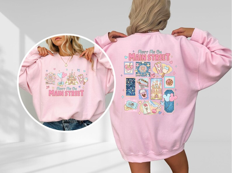 May include: A light pink sweatshirt with the text "Meet Me On Main Street" and various cartoon illustrations. The back of the sweatshirt features additional themed graphics. The sweatshirt is a casual style.