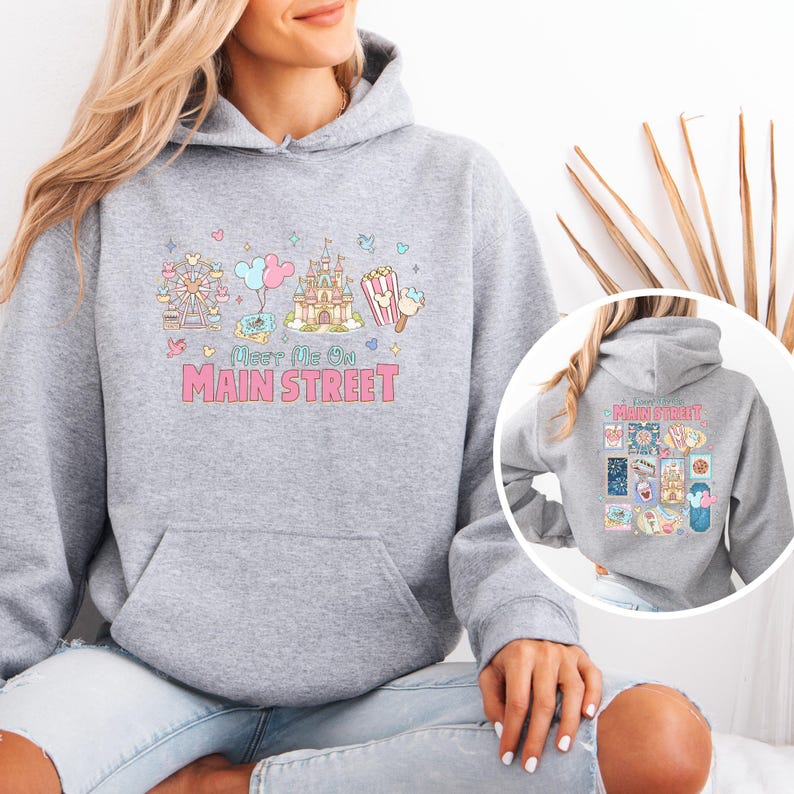 May include: A light gray hoodie with a front pocket and a colorful graphic. The graphic includes a Ferris wheel, balloons, a castle, popcorn, and ice cream, with the text "Meet Me On Main Street" in pink.