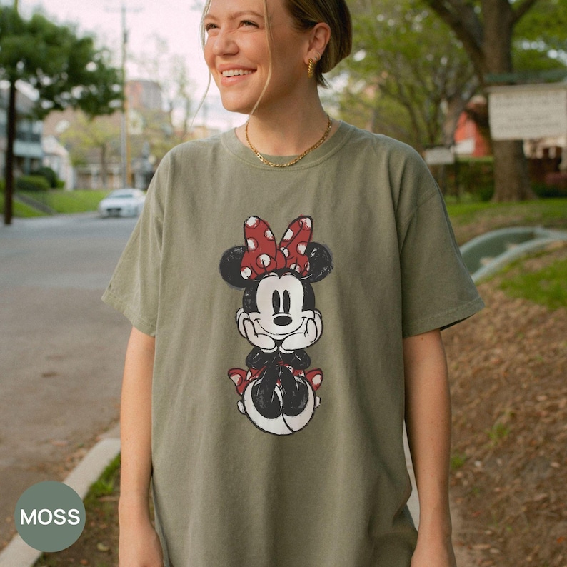 Puede incluir: Camiseta verde oliva con un gr&aacute;fico desgastado de Minnie Mouse. El personaje de dibujos animados tiene un lazo rojo con lunares blancos. La palabra "MOSS" est&aacute; en un c&iacute;rculo en la esquina inferior izquierda. La camiseta la lleva una persona.