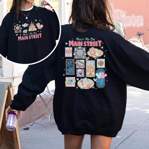 May include: Black sweatshirt with a colorful graphic design on the front and back. The front design says "Meet Me On Main Street" with a castle and other icons. The back has a similar design with stamps.