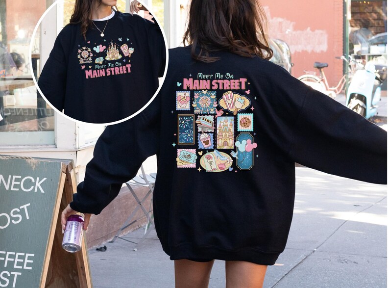 May include: Black sweatshirt with a colorful graphic design on the front and back. The front design says "Meet Me On Main Street" with a castle and other icons. The back has a similar design with stamps.