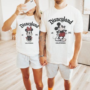 May include: Two people wearing matching white t-shirts with Disneyland graphics. One shirt features Minnie Mouse, the other Mickey Mouse. Both shirts have the text "Disneyland est. 1955 CALIFORNIA" in black.