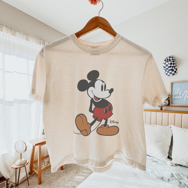 May include: Cream-colored t-shirt featuring a classic Mickey Mouse graphic. The cartoon character is depicted in black, red, and brown, with the Disney logo at the bottom. The shirt is hanging on a wooden hanger.