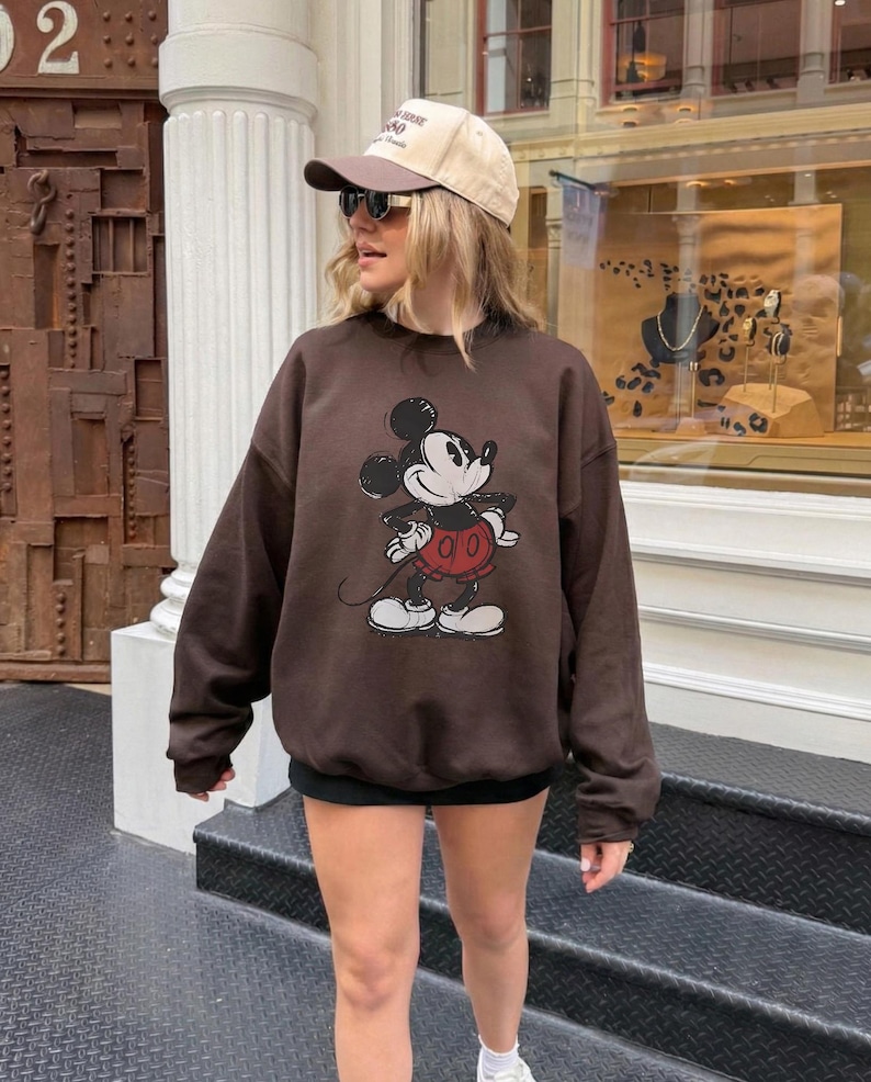 May include: A brown sweatshirt featuring a Mickey Mouse graphic. The sweatshirt has long sleeves and a crew neck. The image also shows a beige baseball cap and sunglasses.