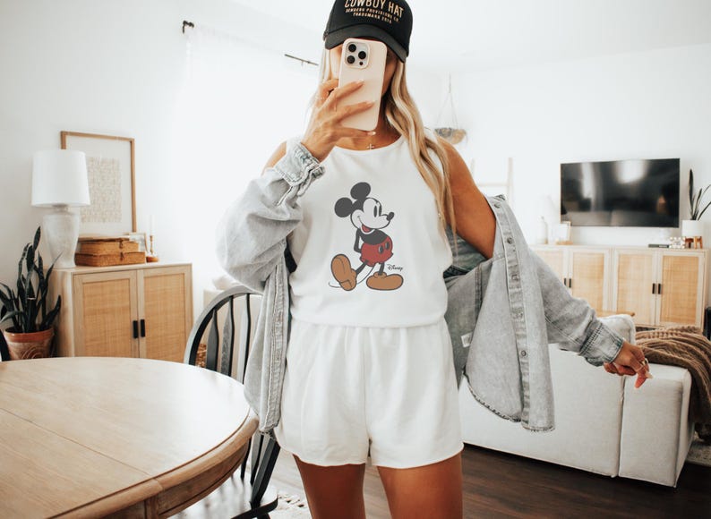 May include: White tank top and shorts set featuring a Mickey Mouse graphic. The outfit is paired with a black baseball cap with white text. A light blue denim jacket is worn over the shoulders.