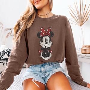 Puede incluir: Sudadera marr&oacute;n con un gr&aacute;fico desgastado de Minnie Mouse con un lazo rojo y lunares. La sudadera es de color marr&oacute;n liso y tiene mangas largas. El gr&aacute;fico est&aacute; centrado en la parte delantera de la prenda.