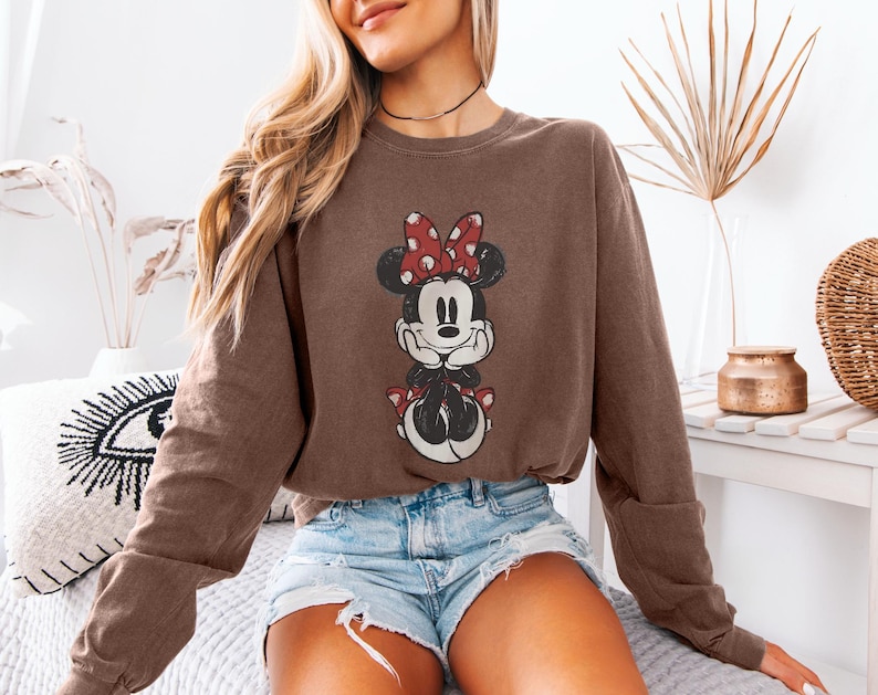 Puede incluir: Sudadera marr&oacute;n con un gr&aacute;fico desgastado de Minnie Mouse con un lazo rojo y lunares. La sudadera es de color marr&oacute;n liso y tiene mangas largas. El gr&aacute;fico est&aacute; centrado en la parte delantera de la prenda.