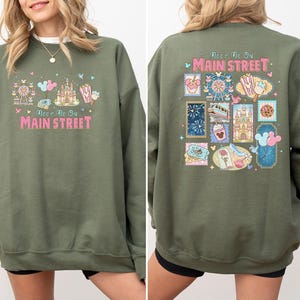 May include: Olive green sweatshirt with the text "Meet Me On Main Street" in pink and white. The front features a castle, Ferris wheel, and popcorn graphic. The back has a stamp-like design with various park icons.