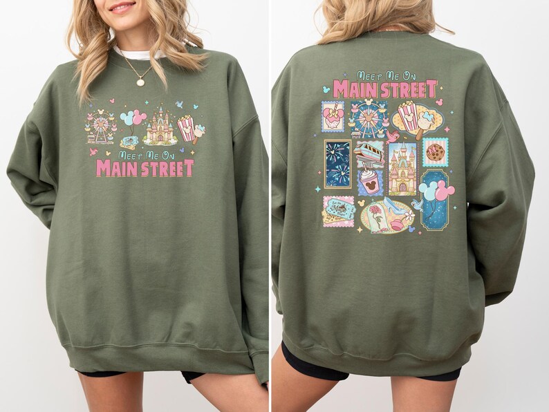 May include: Olive green sweatshirt with the text "Meet Me On Main Street" in pink and white. The front features a castle, Ferris wheel, and popcorn graphic. The back has a stamp-like design with various park icons.