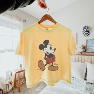 May include: Yellow t-shirt featuring a graphic of Mickey Mouse in black, red, and brown. The Disney character is walking with a smile. The shirt is hanging on a wooden hanger.