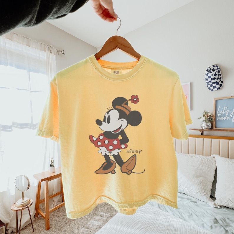 May include: Yellow t-shirt featuring a Minnie Mouse graphic. The graphic shows Minnie Mouse in a red and white polka dot dress, black shoes, and a flower on her head. The Disney logo is printed below the character.
