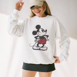 May include: White sweatshirt featuring a distressed graphic of Mickey Mouse in black, red, and white. The sweatshirt has long sleeves and a crew neck. The person is wearing a beige baseball cap and sunglasses.