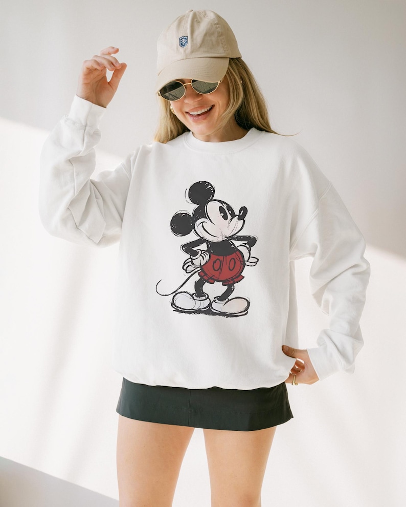 May include: White sweatshirt featuring a distressed graphic of Mickey Mouse in black, red, and white. The sweatshirt has long sleeves and a crew neck. The person is wearing a beige baseball cap and sunglasses.