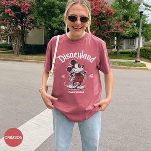 May include: Crimson-colored t-shirt with the Disneyland logo and a graphic of Mickey Mouse. The shirt features the text "est. 1955 CALIFORNIA". The shirt is paired with light blue jeans.