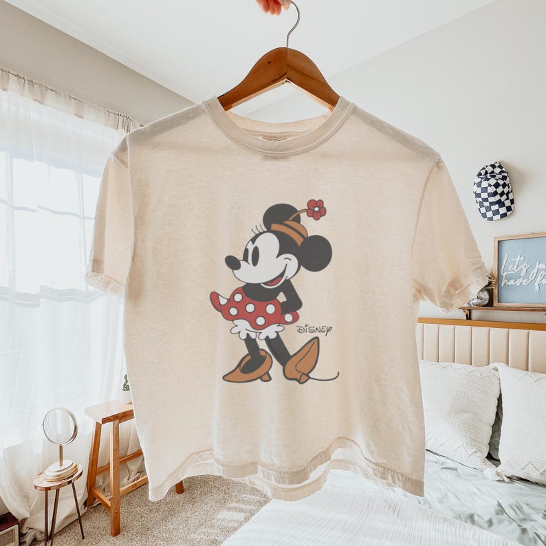 May include: Cream-colored t-shirt featuring a graphic of Minnie Mouse in a red polka-dot dress and matching bow. The Disney logo is printed below the character. The shirt is hanging on a wooden hanger.
