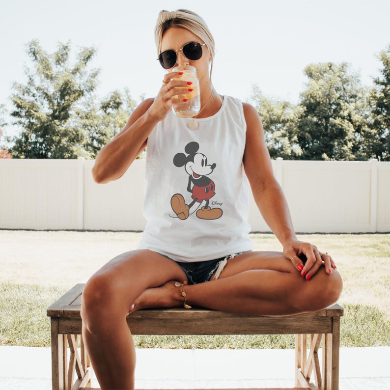 May include: White tank top featuring a classic Mickey Mouse graphic. The cartoon character is depicted in black, red, and tan. The tank top is sleeveless and has a round neckline. The Disney logo is printed below the graphic.
